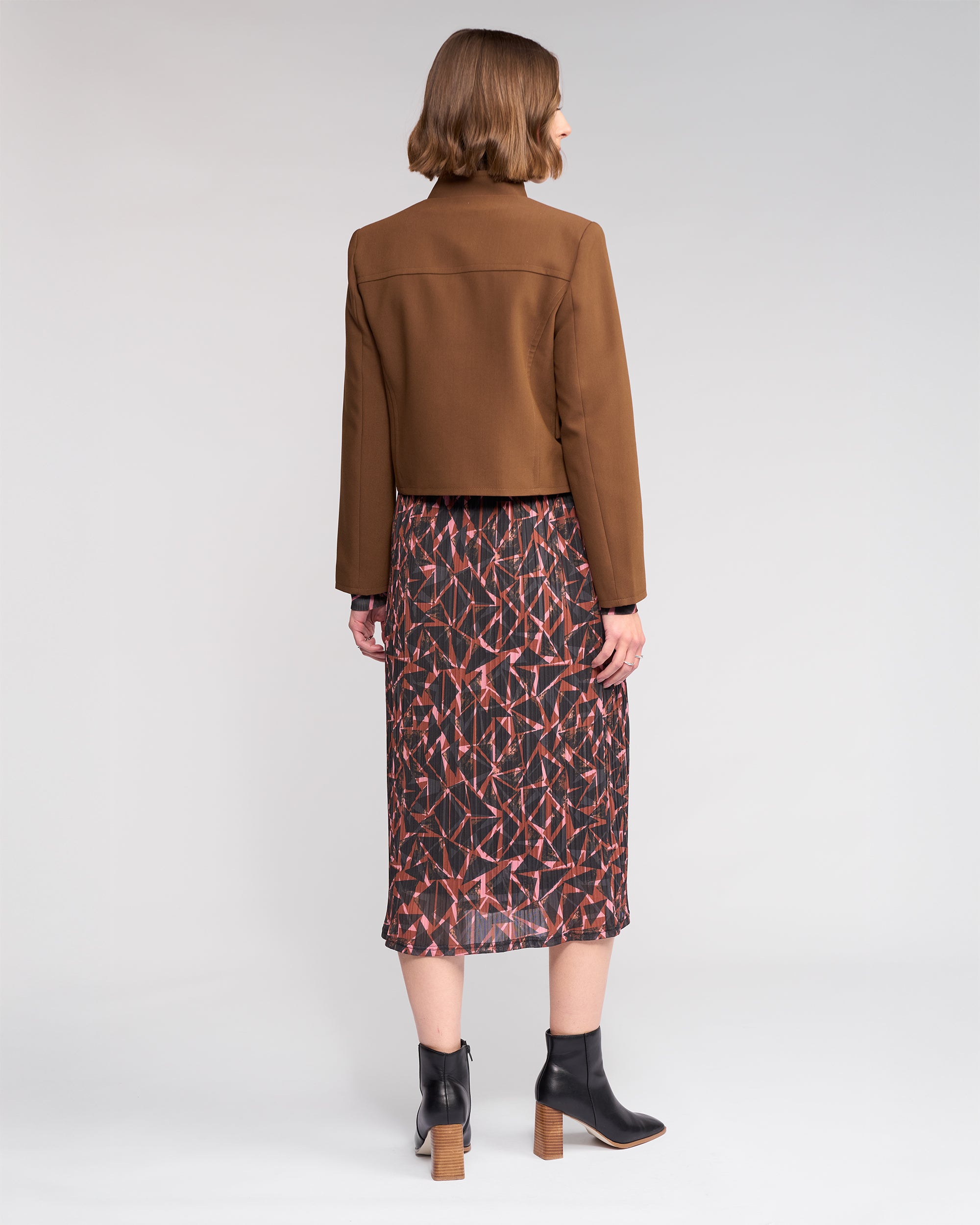 A woman faces away, wearing the Knewe Label Soho Jacket in Cedar with a stand collar, paired with a geometric patterned midi skirt and black ankle boots with wooden heels, set against a plain light background.