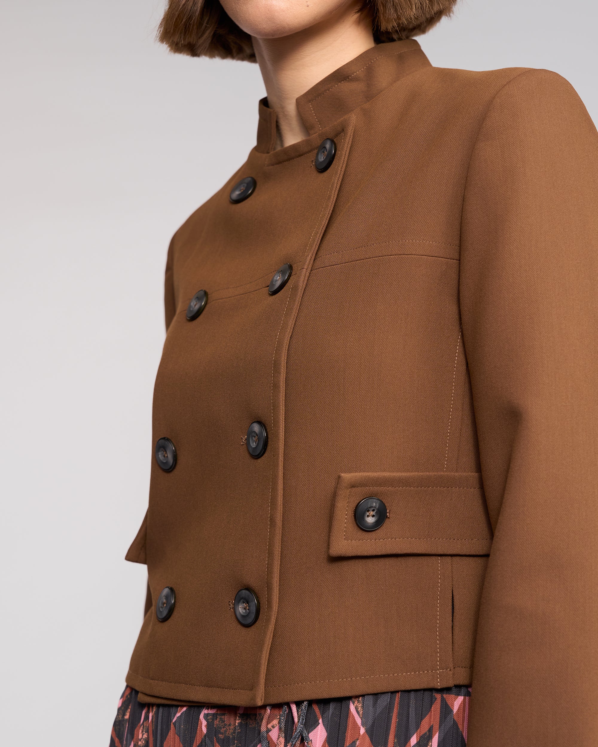 A woman with a short brown bob models the Soho Jacket - Cedar by Knewe Label, featuring a golden brown double-breasted design with black buttons and flap pockets, paired with a patterned skirt. Photo is cropped above her nose against a plain background.