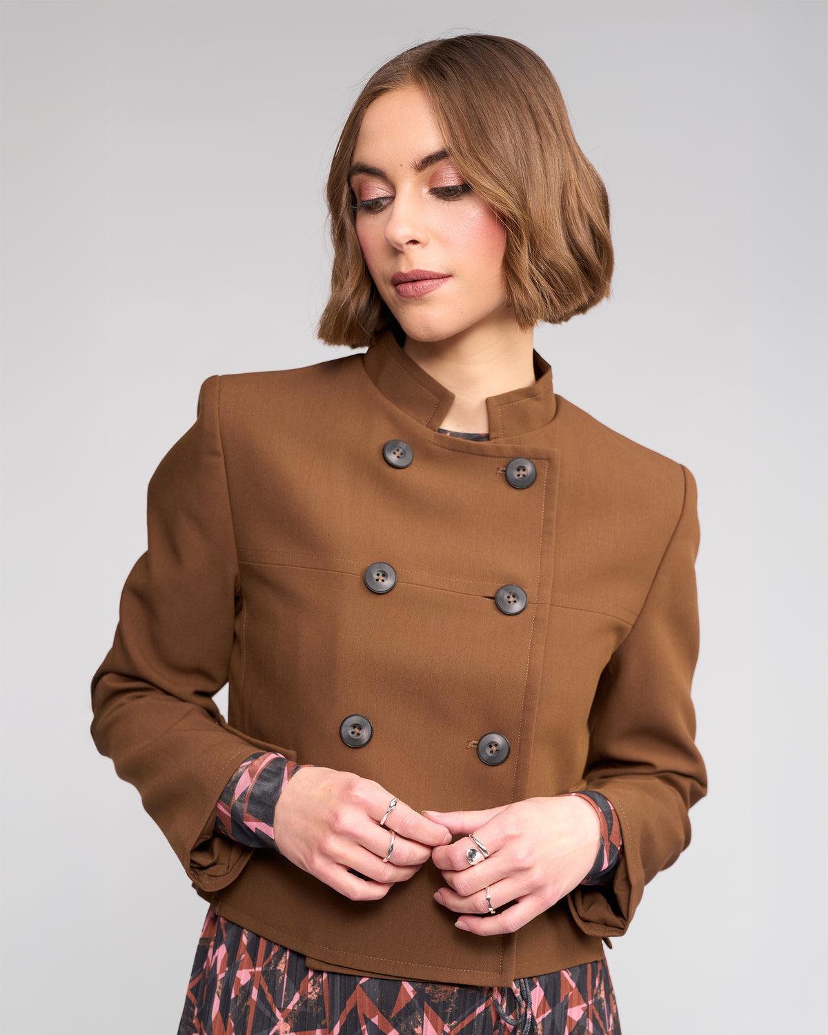 A woman with chin-length brown hair wears the Knewe Label Soho Jacket in Cedar over a patterned dress. She stands against a plain light gray background, looking down with her hands lightly touching in front of her.