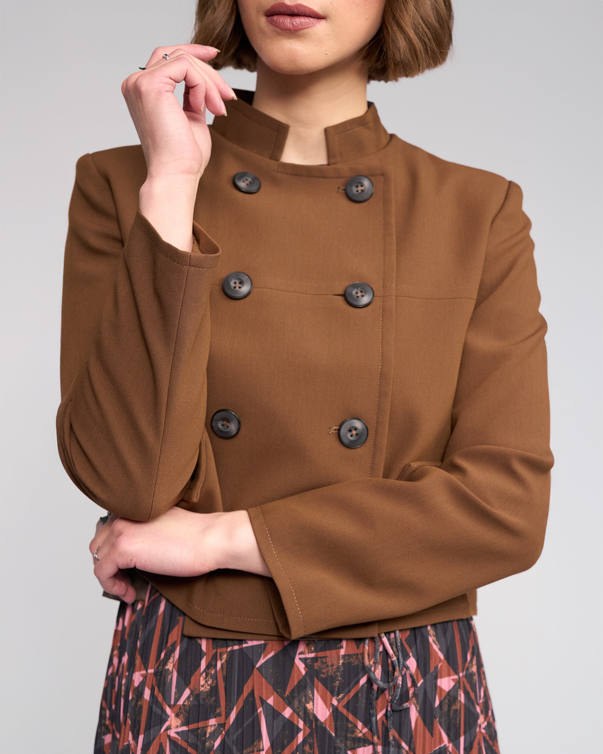 A woman with short brown hair partially covering her features wears the Knewe Label Soho Jacket in Cedar, a golden brown double-breasted style with a stand collar and black buttons, paired with a patterned skirt. She stands with one arm crossed and one hand on her face.