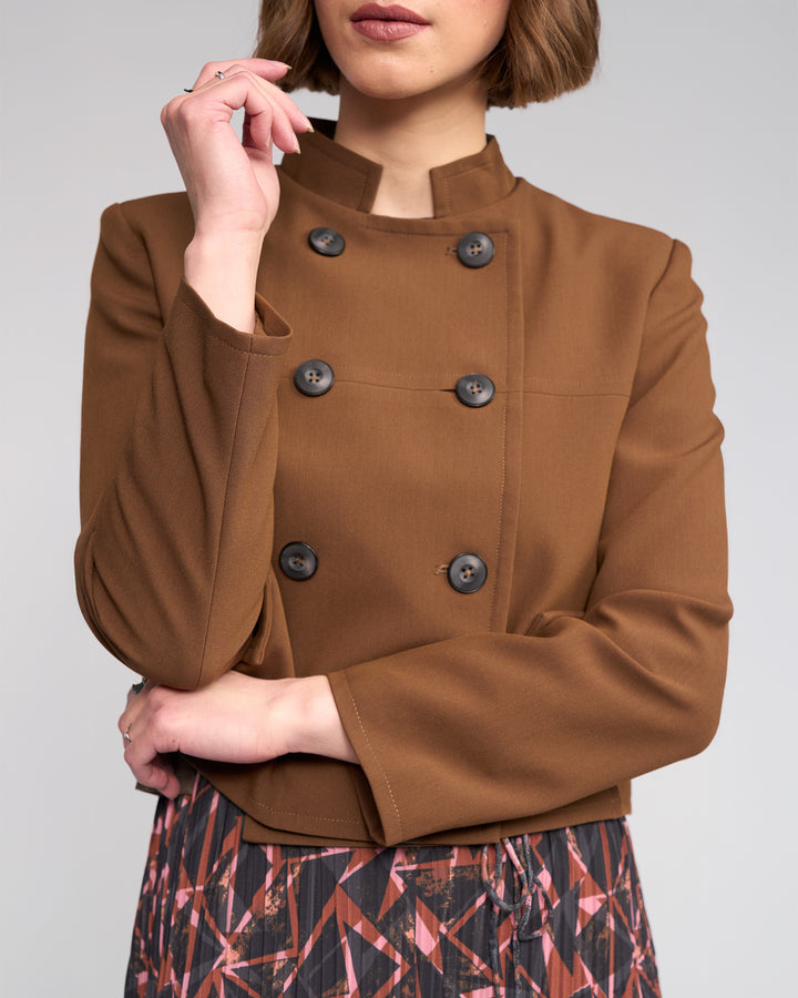 A woman with short brown hair partially covering her features wears the Knewe Label Soho Jacket in Cedar, a golden brown double-breasted style with a stand collar and black buttons, paired with a patterned skirt. She stands with one arm crossed and one hand on her face.