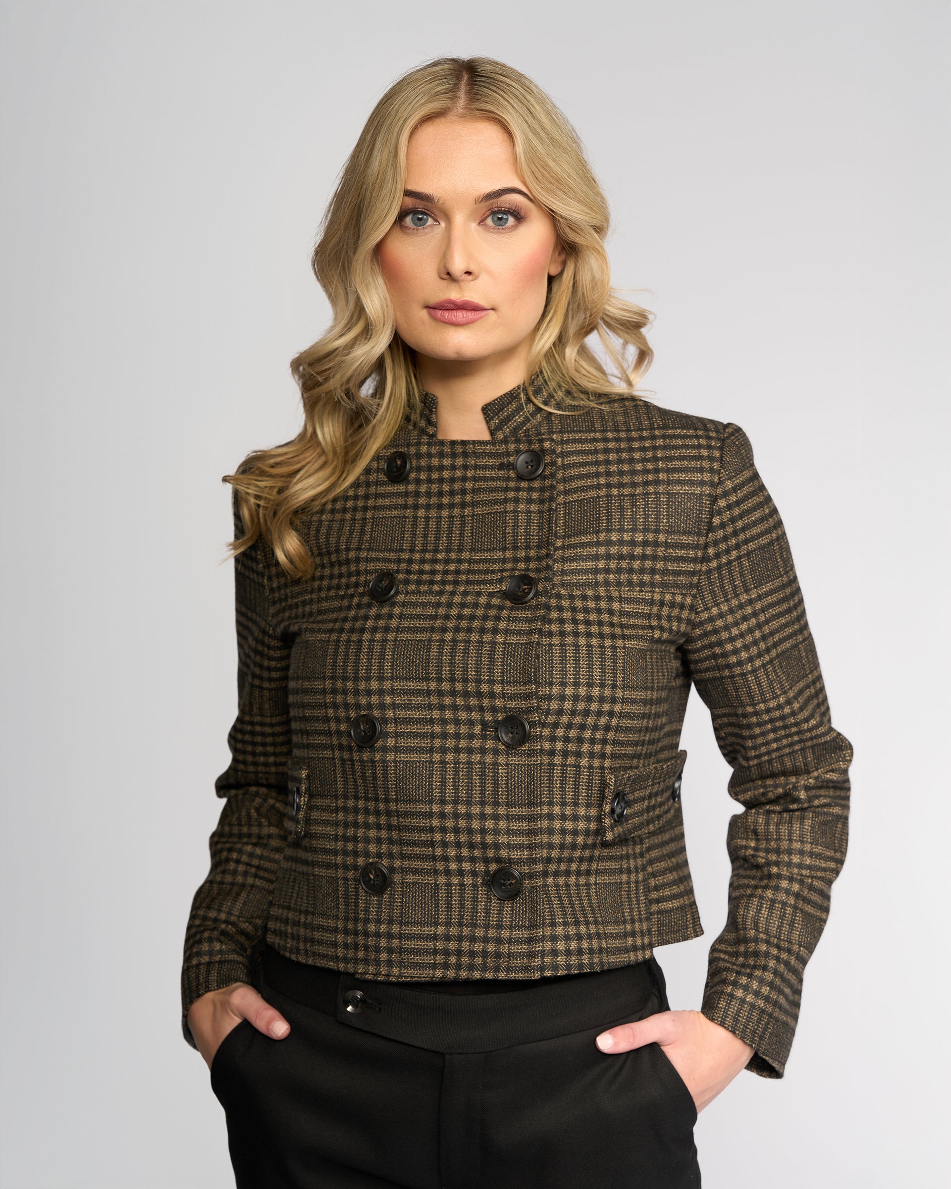 A woman with long blonde hair wears the Knewe Label Soho Jacket - Check, a brown plaid double-breasted winter jacket, and black pants, standing with her hands in her pockets against a plain light background.