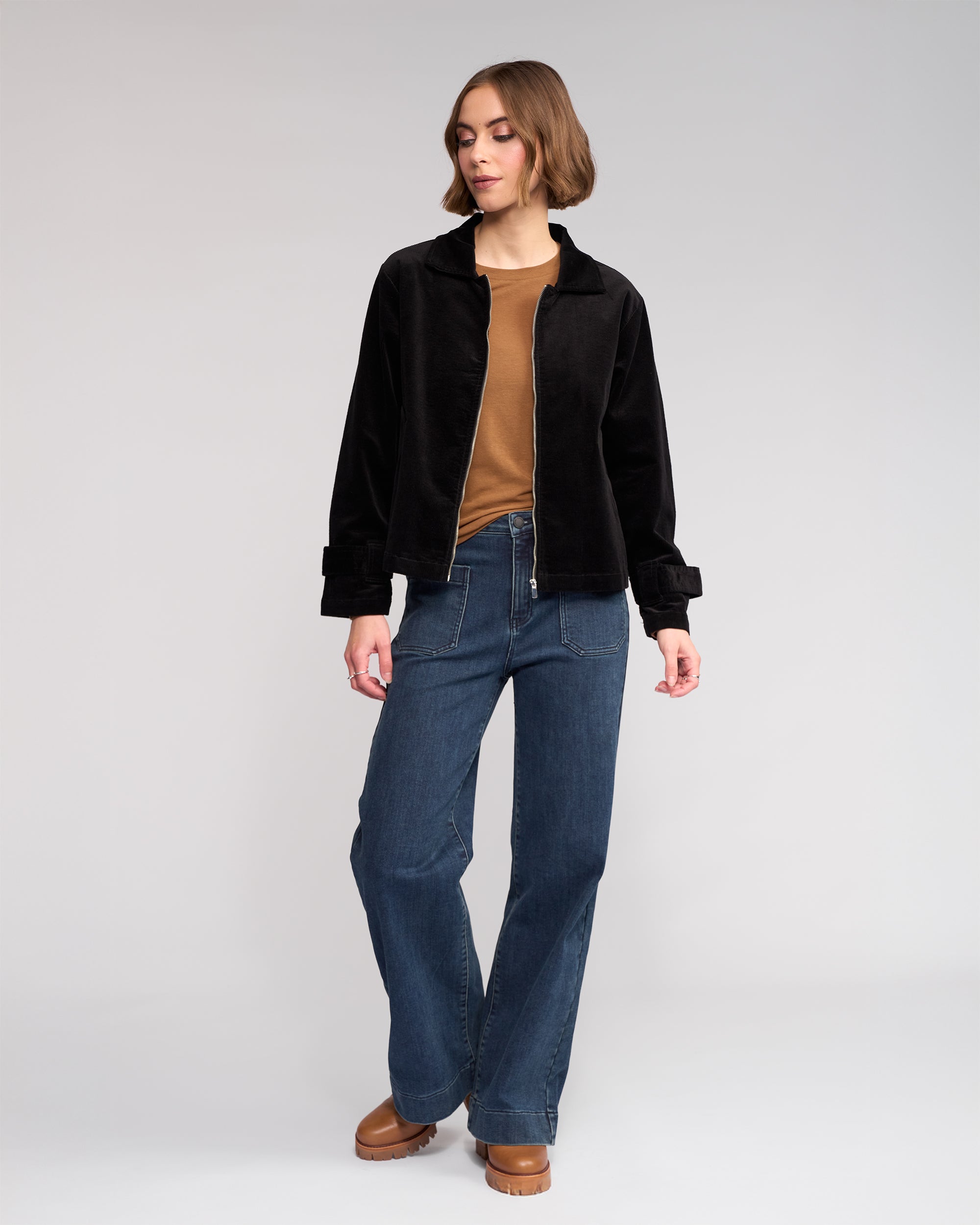 A woman with short brown hair stands against a plain background wearing the Knewe Label Sable Jacket in black, paired with a brown top, blue jeans, and brown platform shoes. She looks to the side with one hand relaxed by her side.
