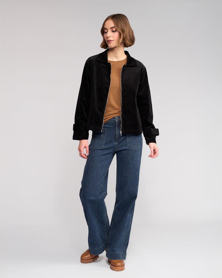 A woman with short brown hair stands against a plain background wearing the Knewe Label Sable Jacket in black, paired with a brown top, blue jeans, and brown platform shoes. She looks to the side with one hand relaxed by her side.
