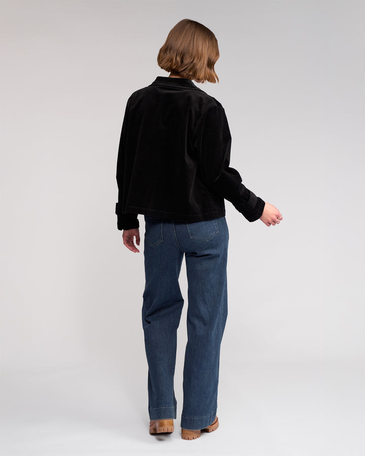 A person with short, light brown hair faces away, wearing the Knewe Label Sable Jacket in black, blue jeans, and brown shoes against a plain white background.