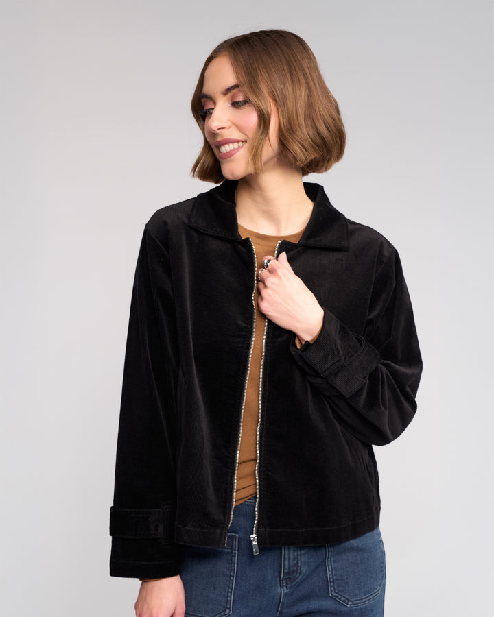 A woman with short brown hair smiles to the side, wearing the Knewe Label Sable Jacket in black over a brown top and blue jeans against a plain light gray background.
