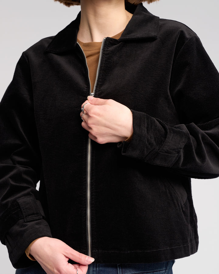 Someone is zipping up the Knewe Label Sable Jacket in black over a brown shirt. Their face is partially out of frame, and the background is plain light gray.