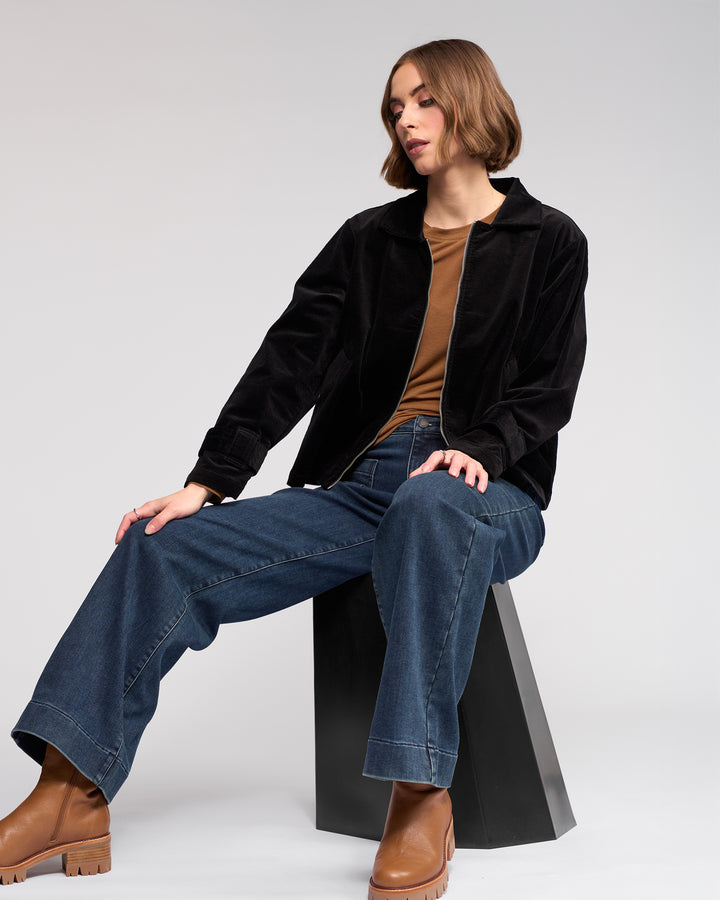 A woman with chin-length brown hair sits on a modern black stool wearing Knewe Label’s Sable Jacket in Black, paired with a brown top, wide-leg blue jeans, and brown heeled boots against a plain light background.