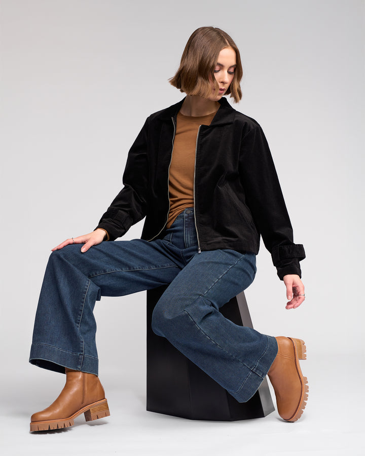 A person with short hair sits sideways on a geometric black stool, wearing Knewe Label's Sable Jacket in Black, a brown top, wide-leg blue jeans, and tan platform boots, looking down with one leg crossed over the other.