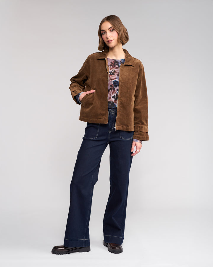 A woman stands against a plain background wearing the Knewe Label Sable Jacket in tan with her hand in her pocket, styled with a patterned top, wide-leg dark blue jeans, and black shoes as she looks confidently at the camera.