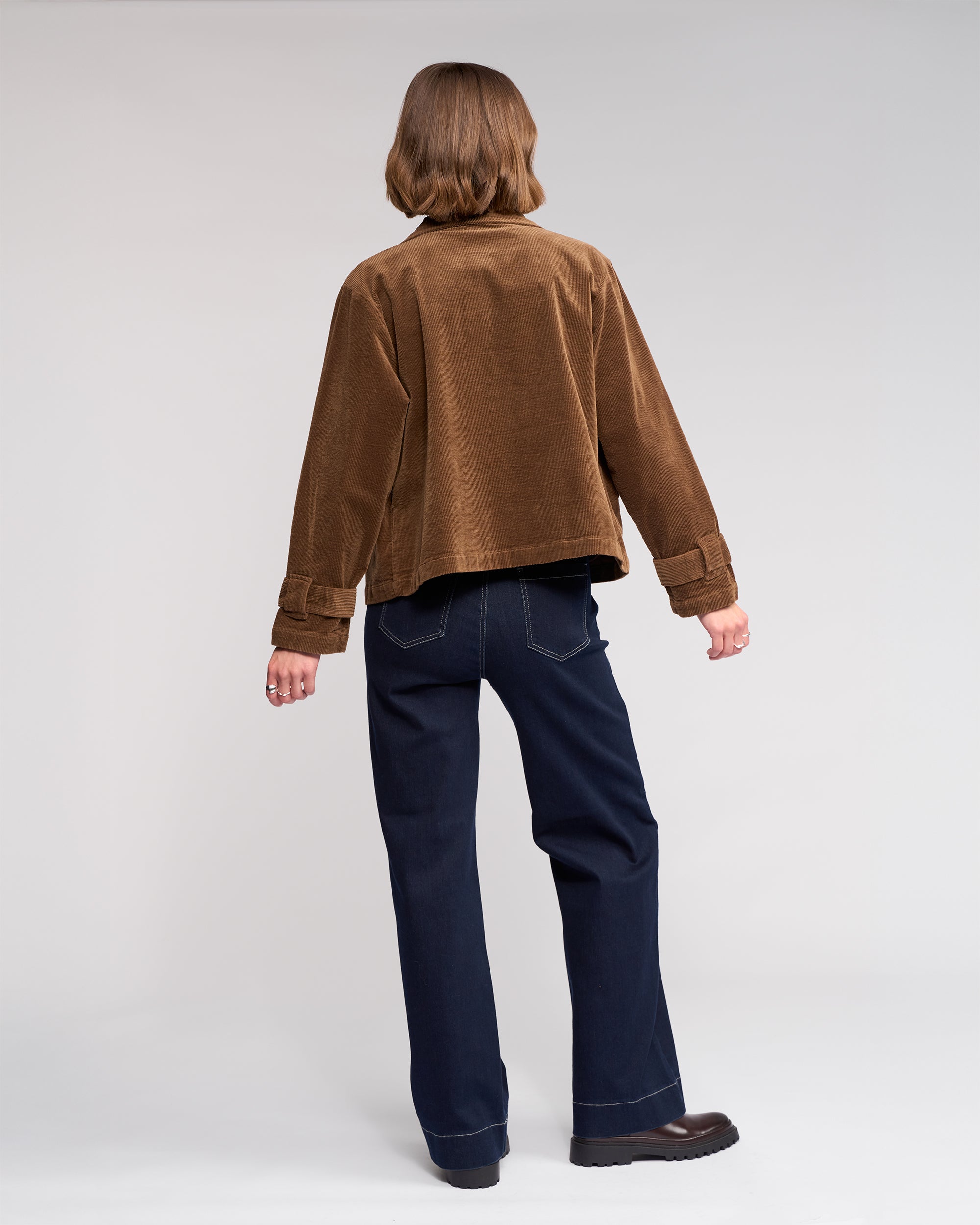 A person with short brown hair is seen from behind wearing the Knewe Label Sable Jacket in tan, paired with dark blue jeans and brown shoes, standing against a plain light background.