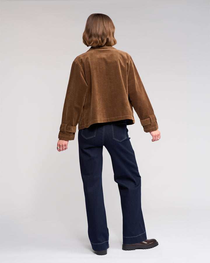 A person with short brown hair is seen from behind wearing the Knewe Label Sable Jacket in tan, paired with dark blue jeans and brown shoes, standing against a plain light background.