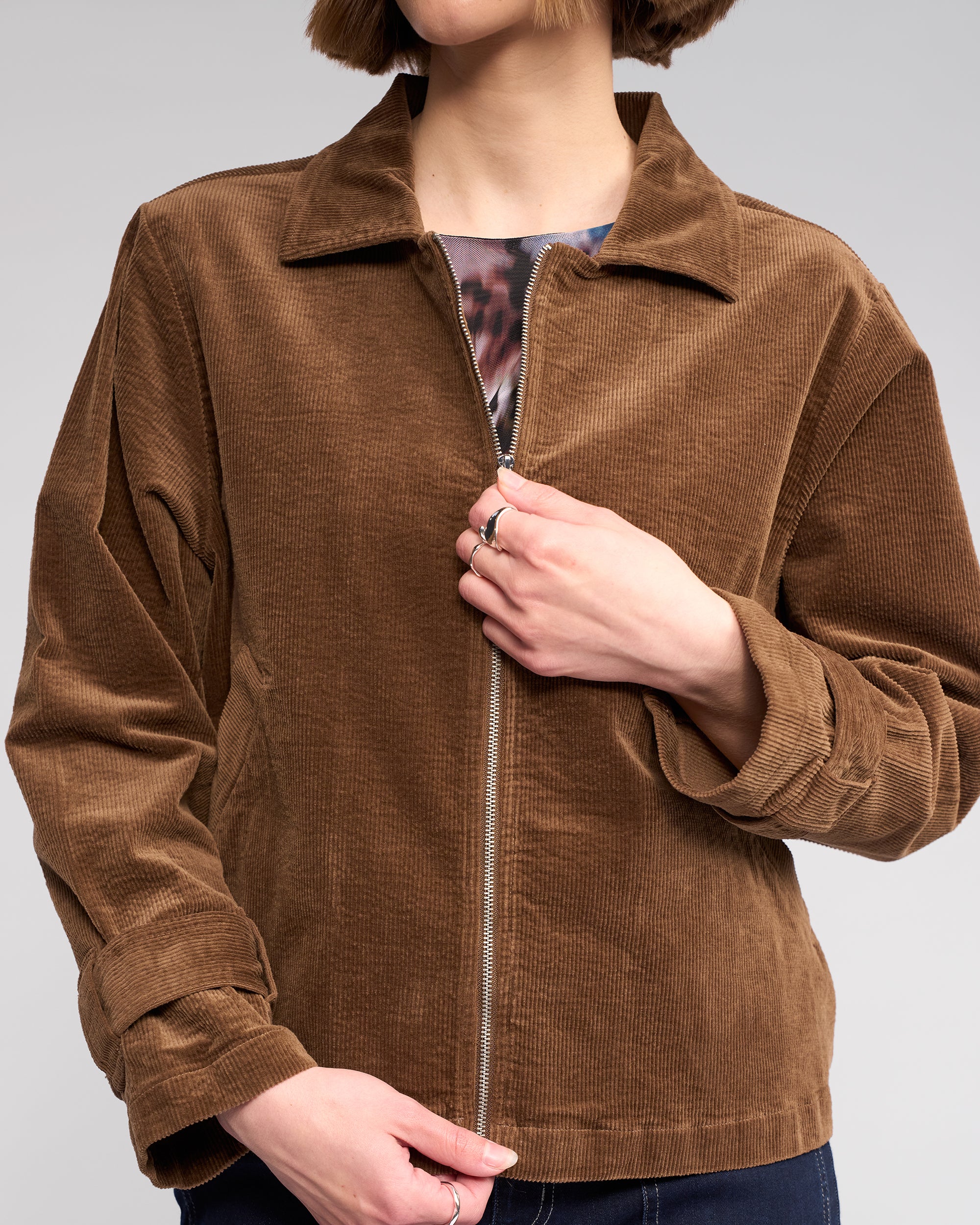 A person with a bob haircut is partially unzipping the Knewe Label Sable Jacket in tan, revealing a patterned shirt underneath. Their face is partly out of frame.