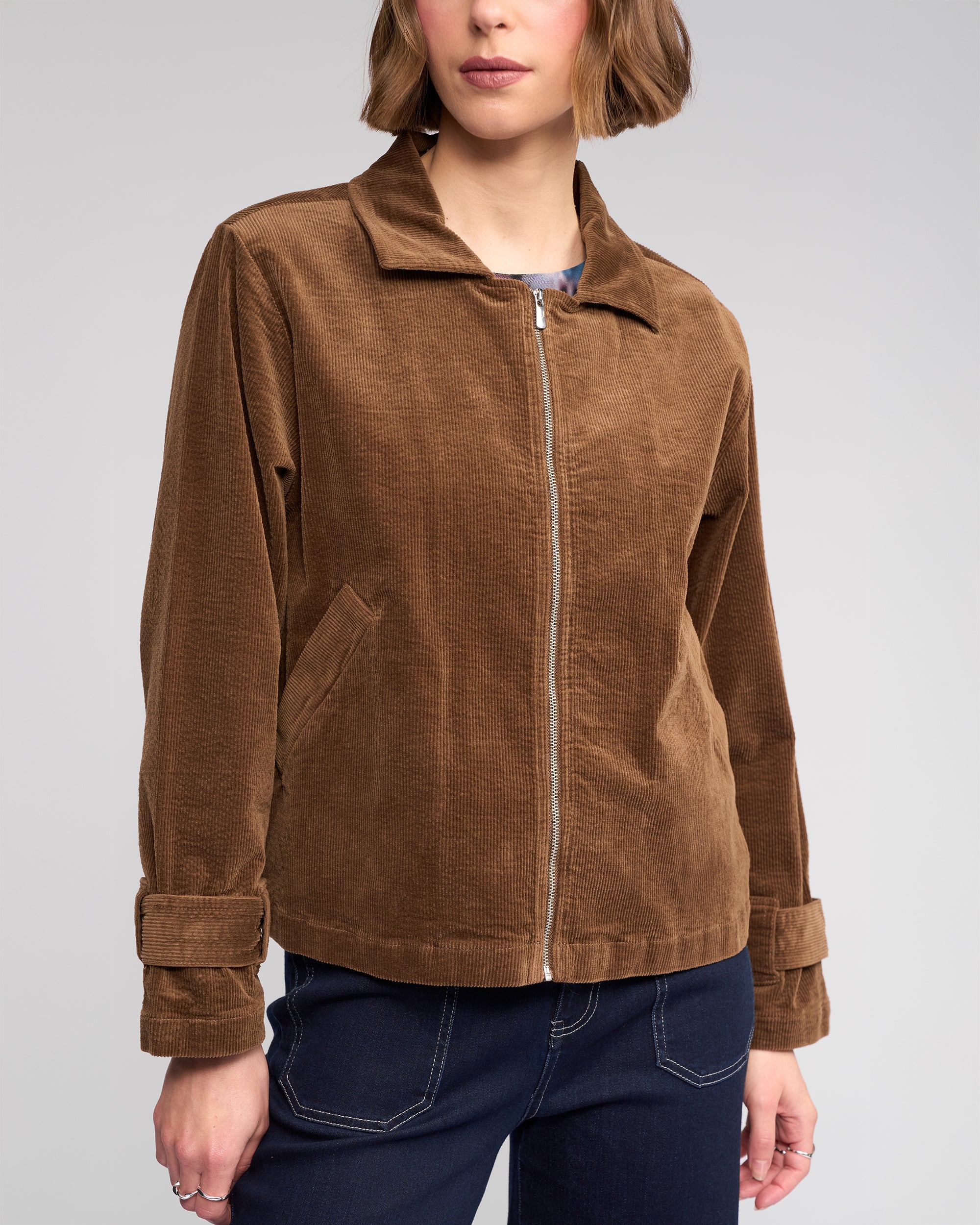 A person wearing the Knewe Label Sable Jacket in tan—a relaxed-fit corduroy jacket with a zip-through front, collar, adjustable cuffs, and side pockets—paired with dark blue jeans against a plain background.