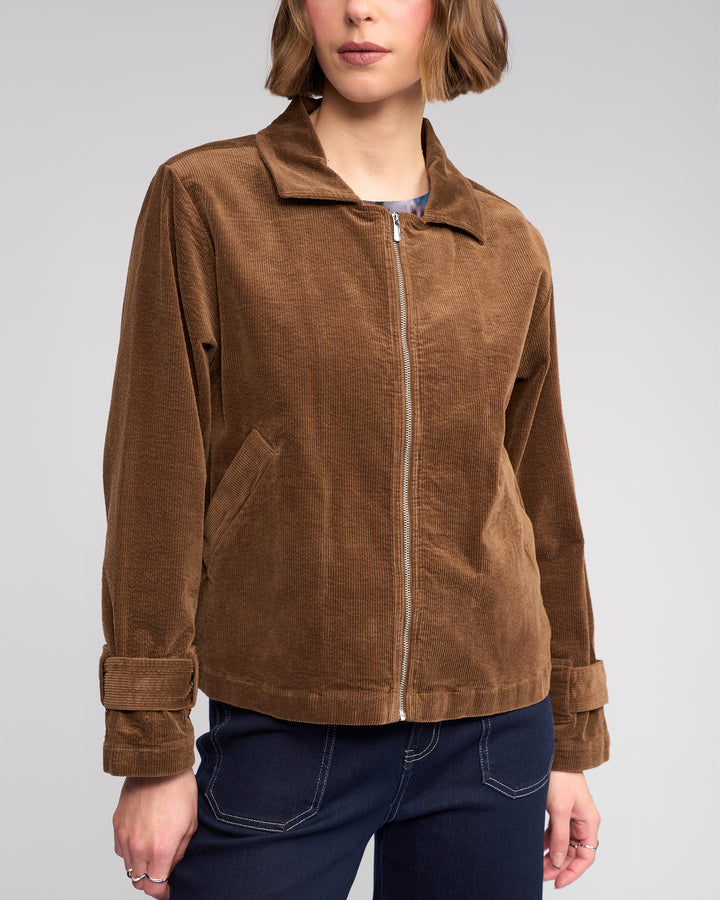 A person wearing the Knewe Label Sable Jacket in tan—a relaxed-fit corduroy jacket with a zip-through front, collar, adjustable cuffs, and side pockets—paired with dark blue jeans against a plain background.