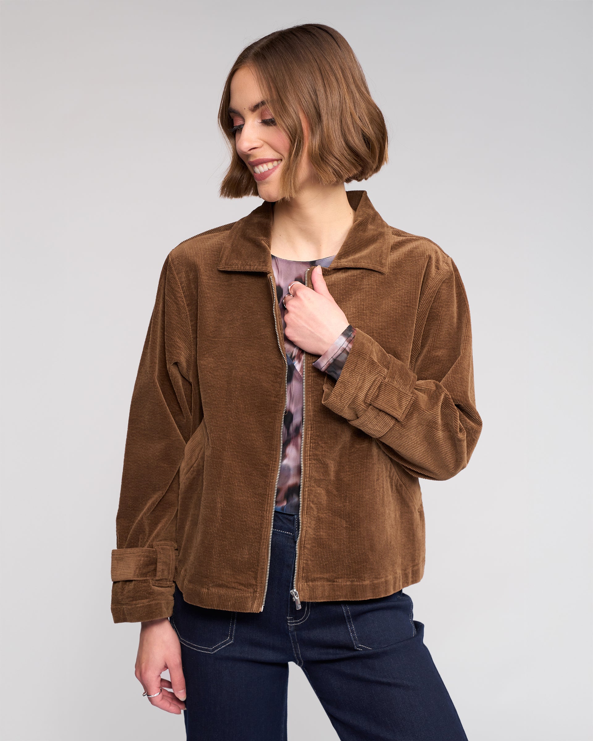 A woman with a short brown bob smiles, standing in the Knewe Label Sable Jacket - Tan, styled over a patterned shirt and dark jeans, her head turned slightly to the side.