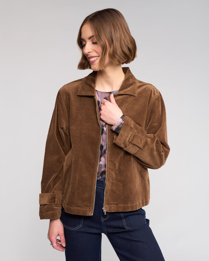 A woman with a short brown bob smiles, standing in the Knewe Label Sable Jacket - Tan, styled over a patterned shirt and dark jeans, her head turned slightly to the side.