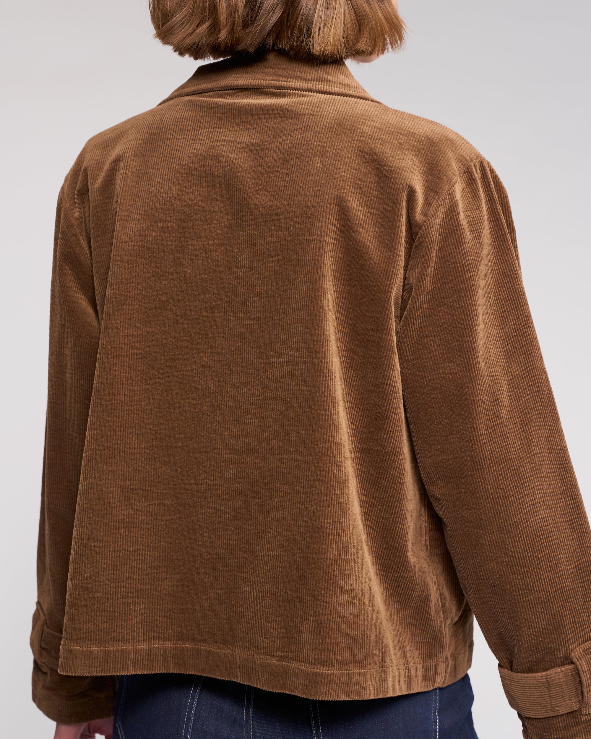 A person with short, light brown hair is shown from the back, wearing the Knewe Label Sable Jacket in tan—a relaxed-fit corduroy style with a zip-through front—paired with dark blue jeans against a plain, neutral background.