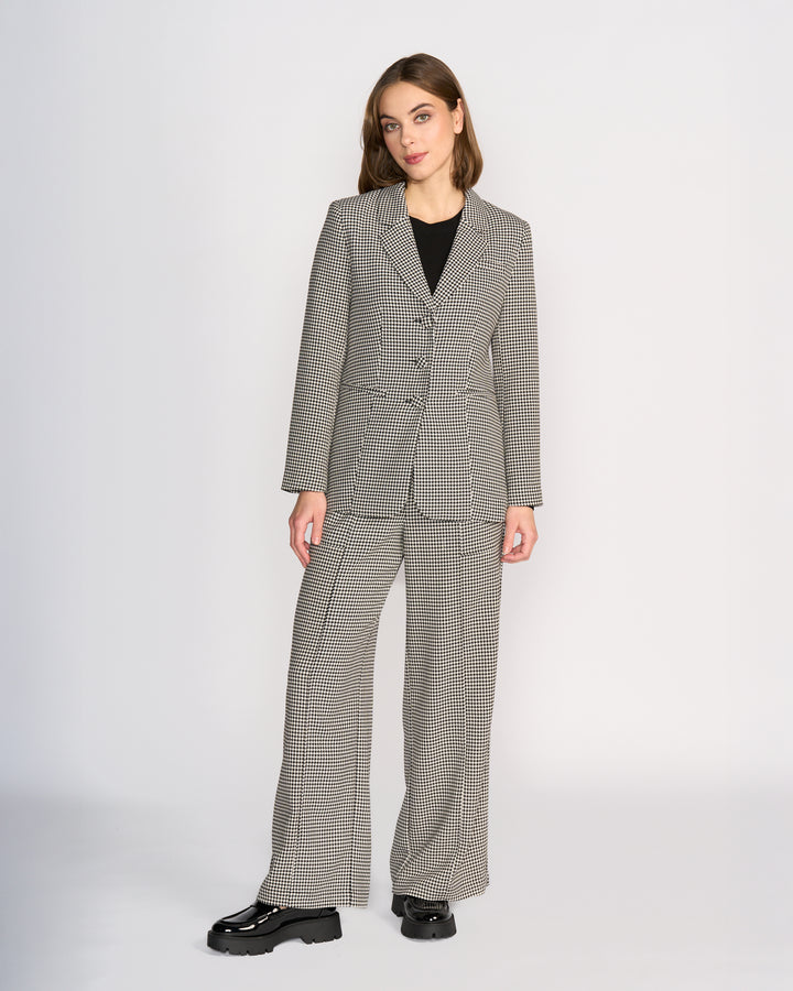 A woman with straight brown hair and a neutral expression stands against a plain light background, wearing Knewe Label's Ryder Blazer - Summit over a black top, paired with matching wide-leg pants and chunky shoes.