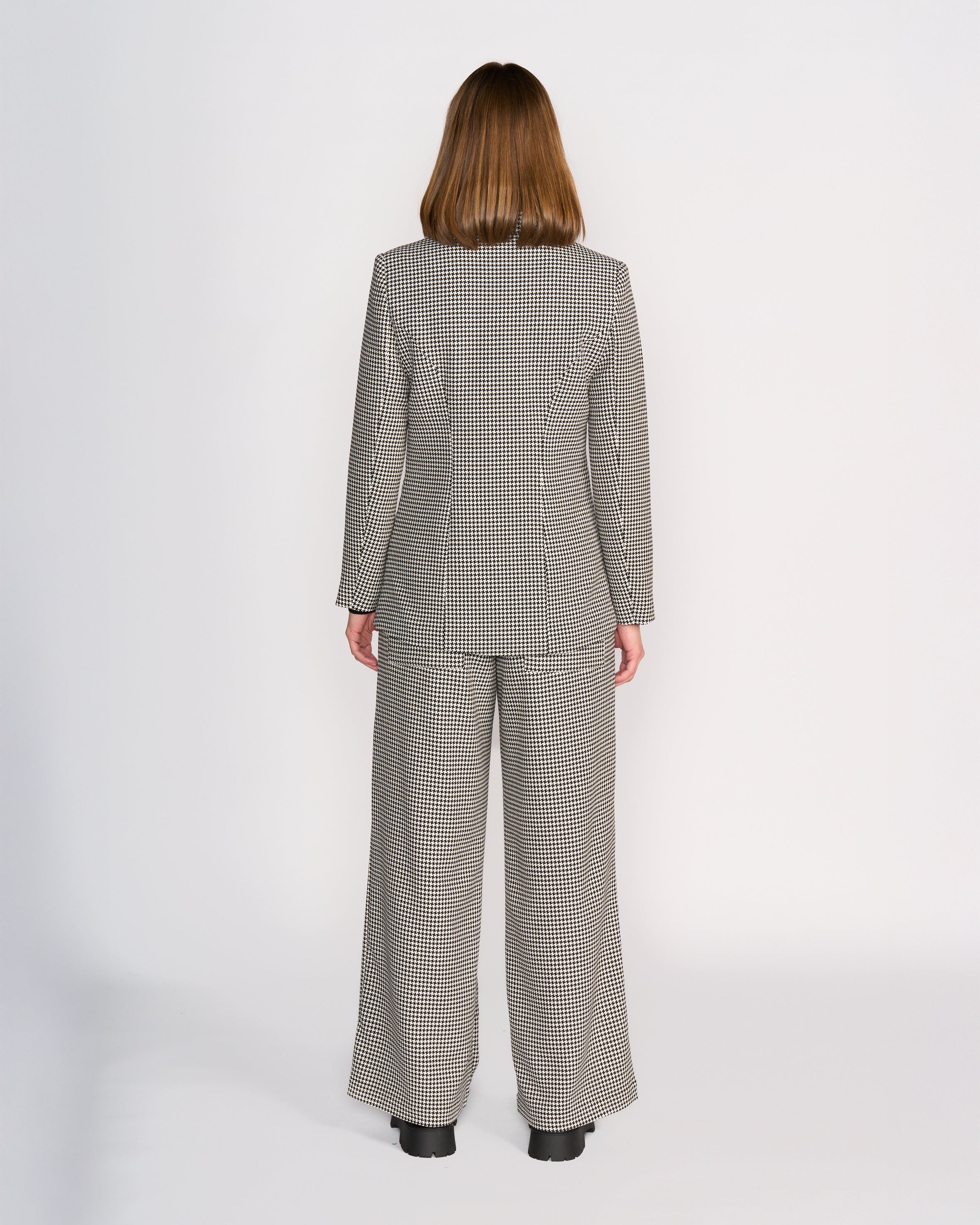 A person with shoulder-length brown hair faces away, wearing the Knewe Label Ryder Blazer - Summit and wide-leg pants in a houndstooth check, contrasted against a plain white background.