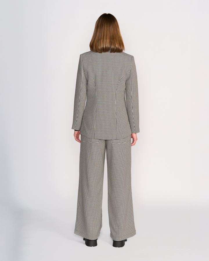 A person with shoulder-length brown hair faces away, wearing the Knewe Label Ryder Blazer - Summit and wide-leg pants in a houndstooth check, contrasted against a plain white background.
