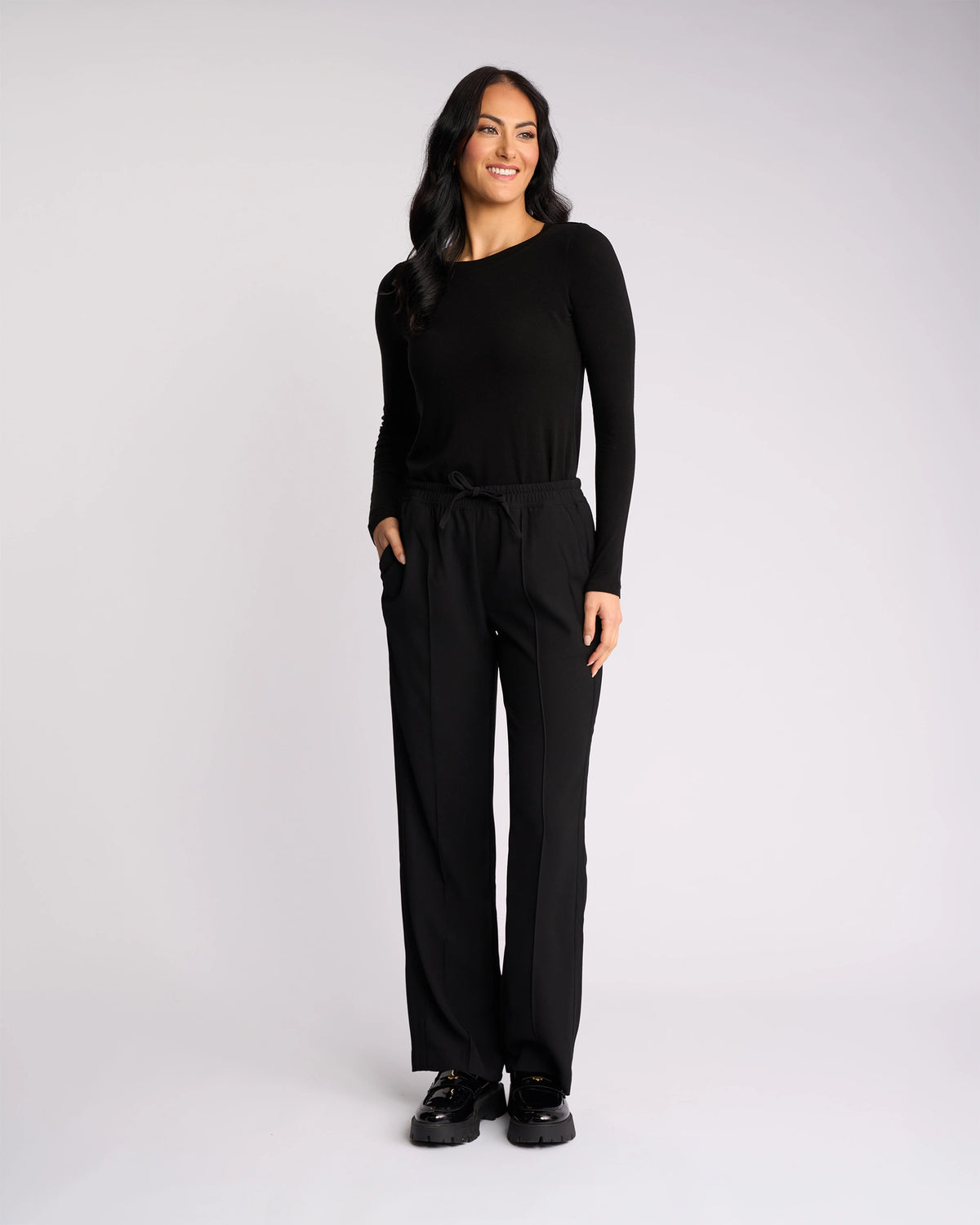 A woman with long dark hair stands against a plain light background, wearing the Bowie Pant - Black/Black from Knewe Label with an elasticated waistband, paired with a black long-sleeve top and shoes, hands in pockets and a slight smile.