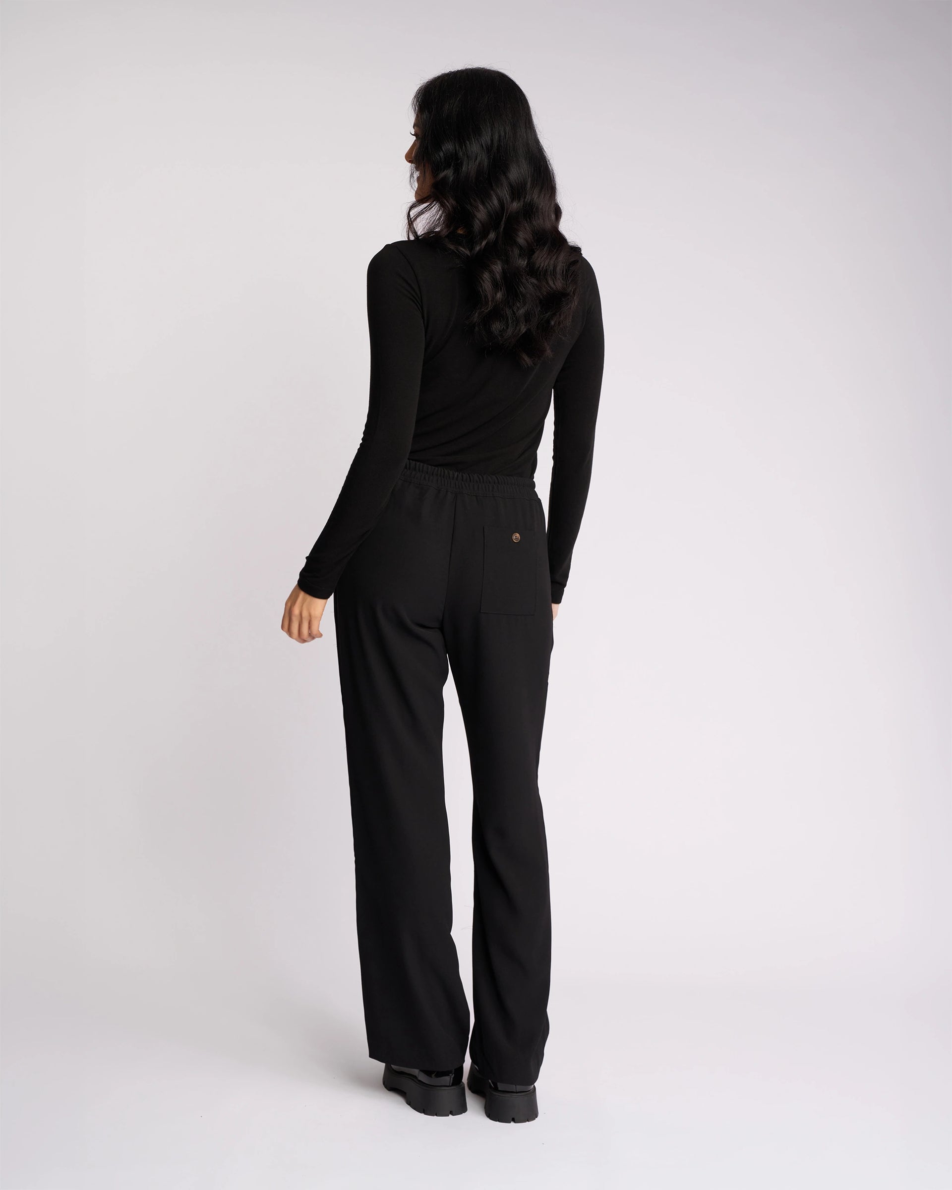 A woman with long dark hair stands facing away, wearing Knewe Label's Bowie Pant - Black/Black, featuring a loose straight-leg fit and elasticated waistband, paired with a fitted black top against a plain white background.