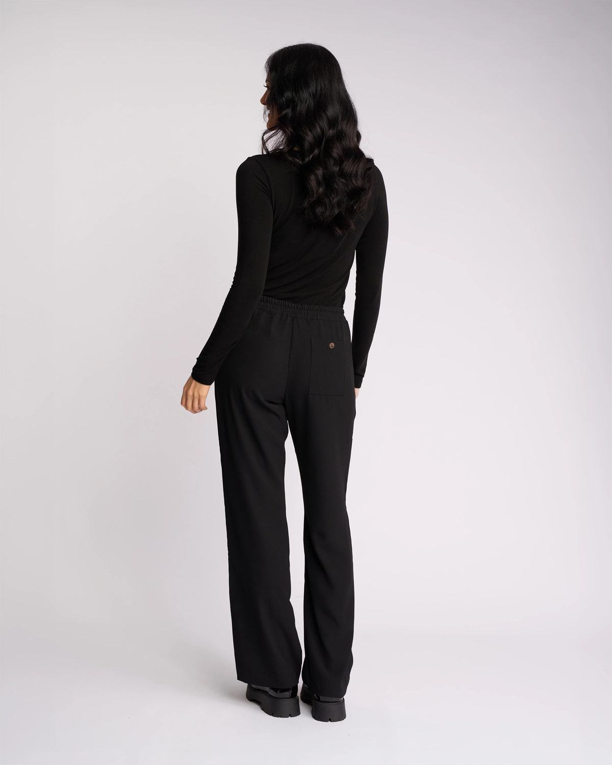A woman with long dark hair stands facing away, wearing Knewe Label's Bowie Pant - Black/Black, featuring a loose straight-leg fit and elasticated waistband, paired with a fitted black top against a plain white background.