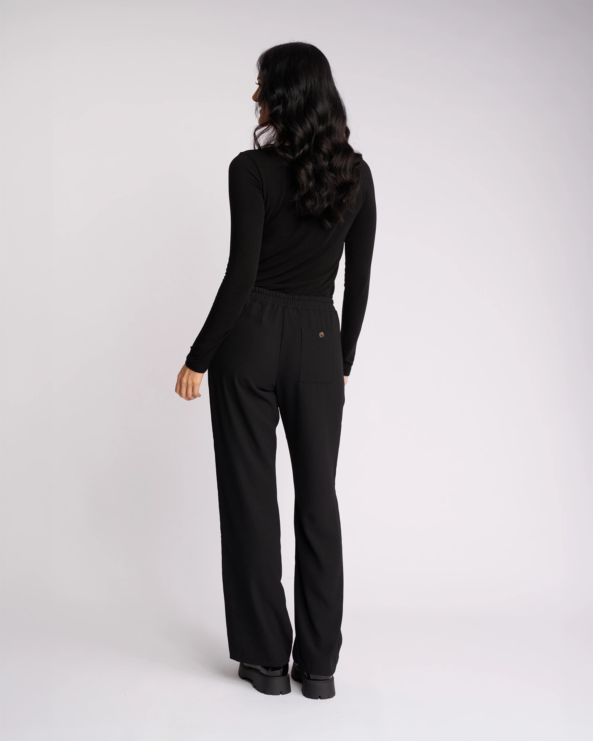 A woman with long dark hair stands facing away, wearing Knewe Label's Bowie Pant - Black/Black, featuring a loose straight-leg fit and elasticated waistband, paired with a fitted black top against a plain white background.