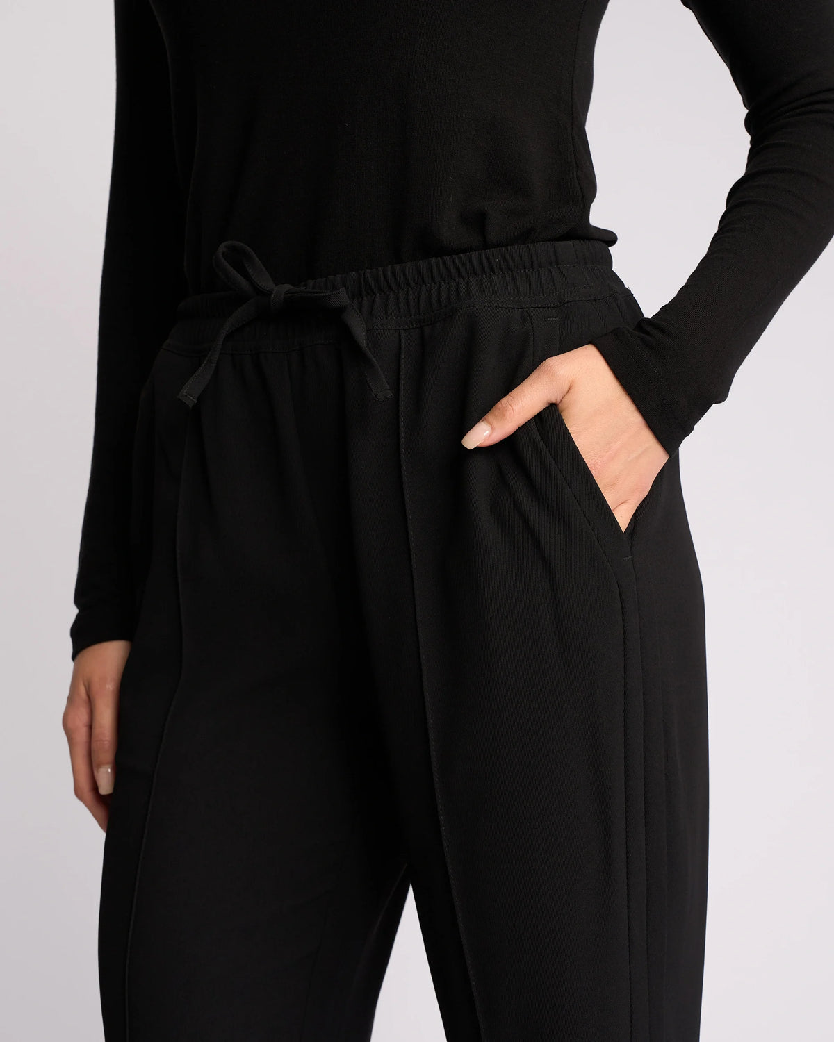 A person models the Knewe Label Bowie Pant - Black/Black, featuring a straight-leg cut and elasticated waistband, with one hand in their pocket against a light background. Only the torso and upper legs are visible.