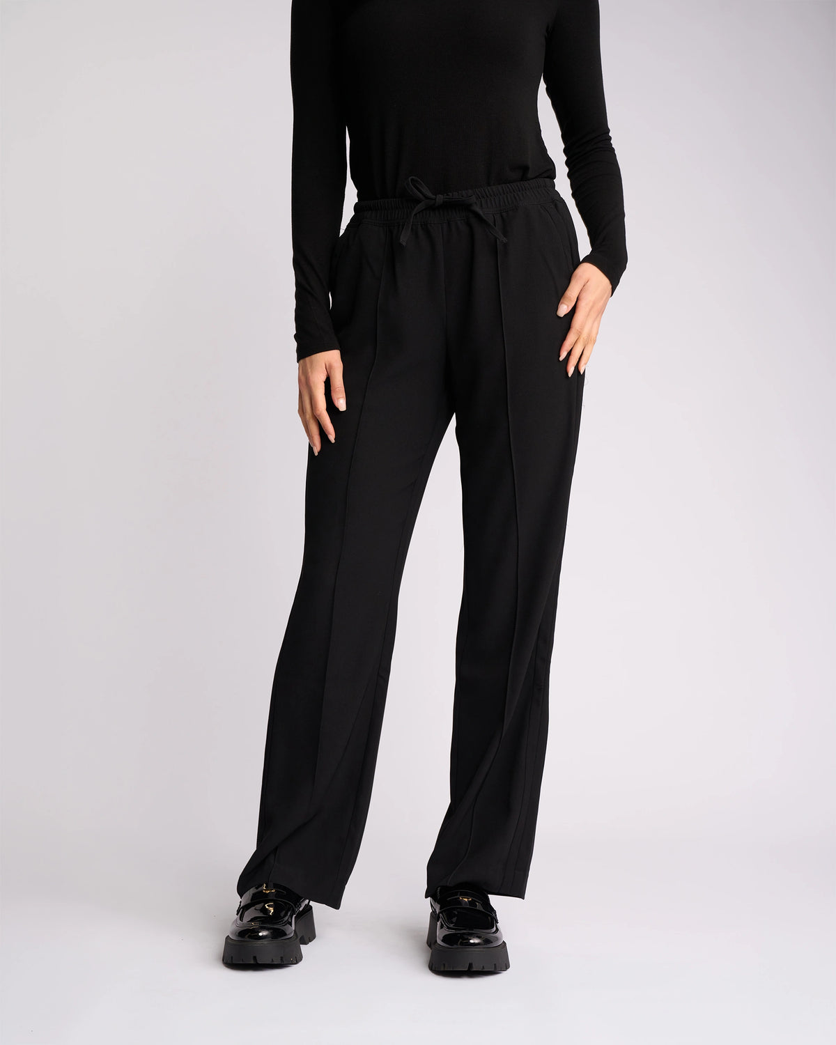 A person models the Knewe Label Bowie Pant - Black/Black, featuring a straight-leg cut and elasticated waistband, paired with a black long-sleeve top and black shoes against a plain light background. The individual's head is not visible.