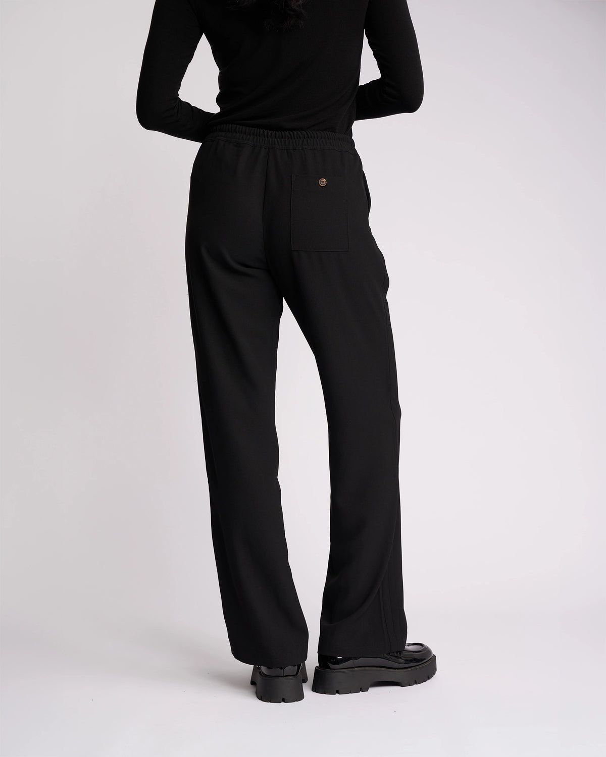 A person stands facing away against a plain white background, wearing the Knewe Label Bowie Pant - Black/Black, paired with a black long-sleeve top and black platform shoes.