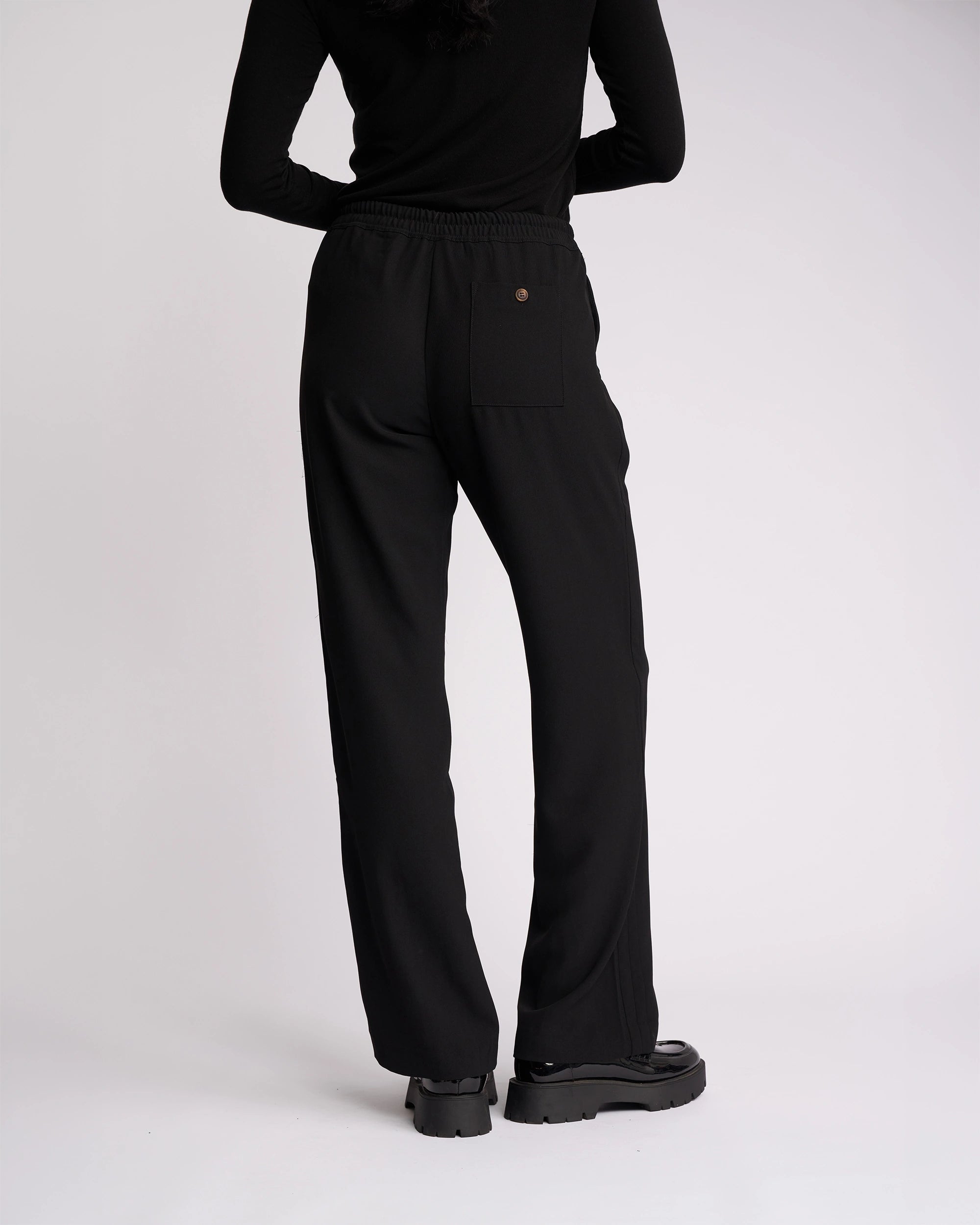 A person stands facing away against a plain white background, wearing the Knewe Label Bowie Pant - Black/Black, paired with a black long-sleeve top and black platform shoes.