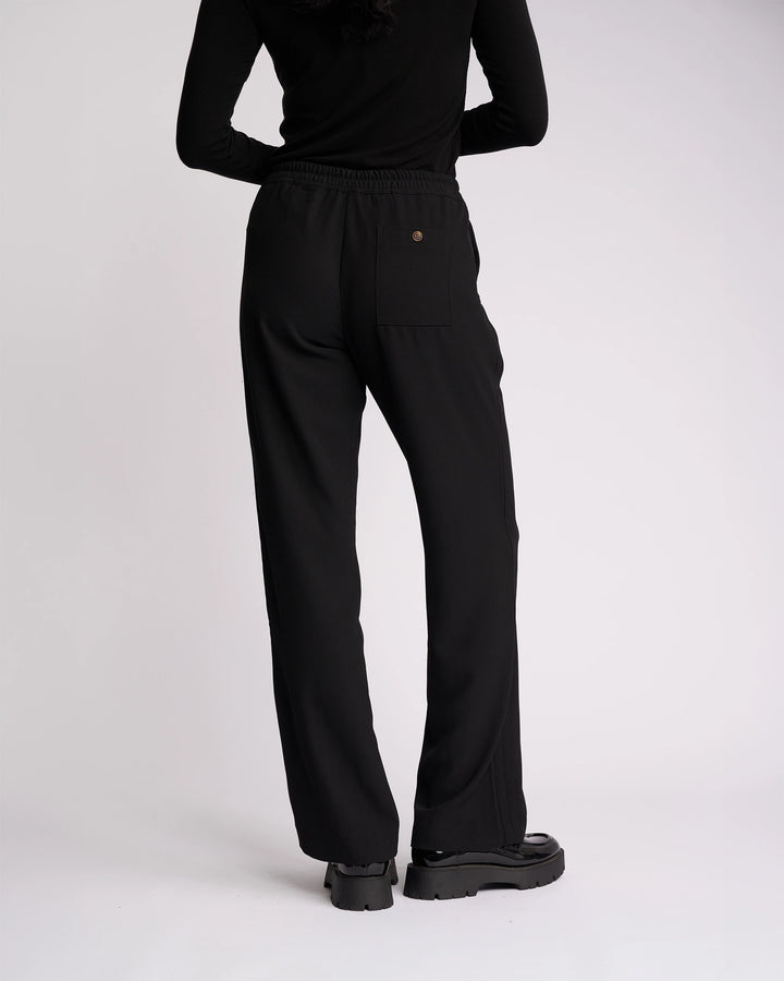 A person stands facing away against a plain white background, wearing the Knewe Label Bowie Pant - Black/Black, paired with a black long-sleeve top and black platform shoes.