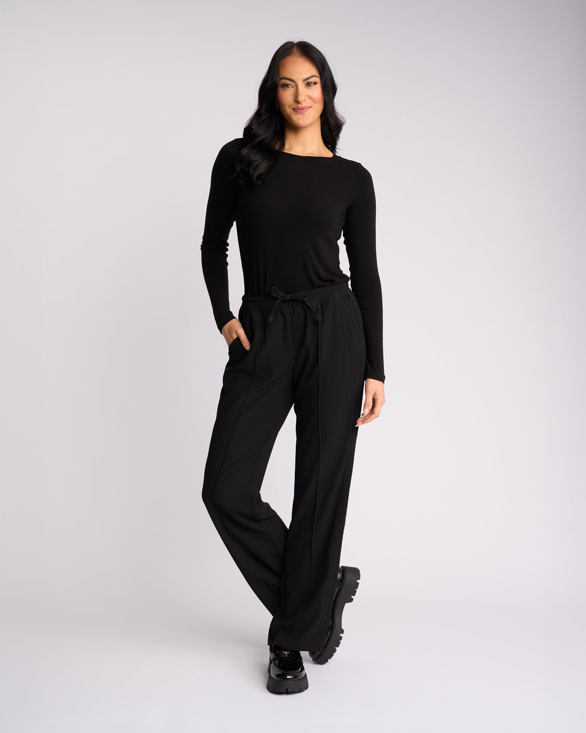 A woman with long dark hair wears the Knewe Label Bowie Pant - Black/Black, paired with a fitted black long-sleeve top and platform shoes. She stands confidently with one hand in her pocket against a plain white background.