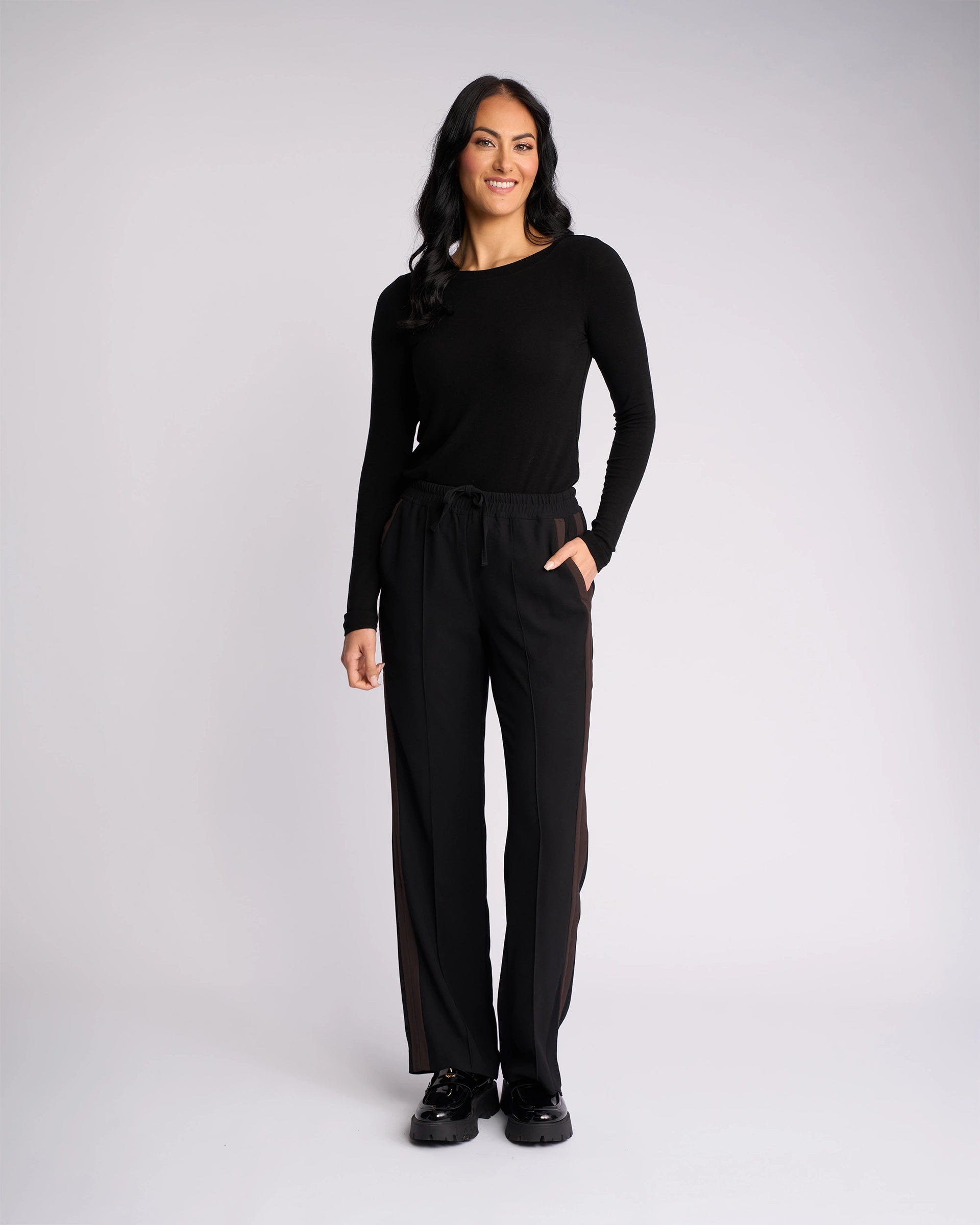 A woman stands against a plain white background, smiling at the camera with her right hand in her pocket. She wears the Knewe Label Bowie Pant in Black/Chocolate, featuring a straight-leg cut and an elasticated waistband.