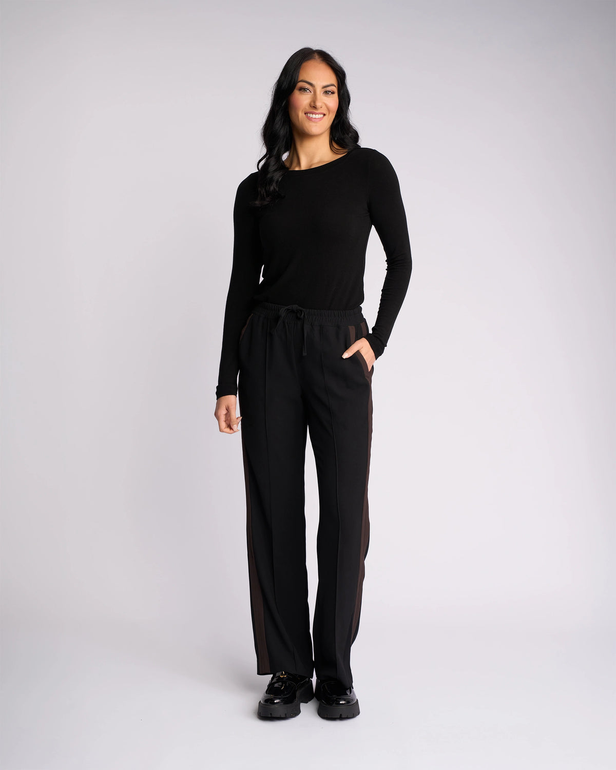 A woman stands against a plain white background, smiling at the camera with her right hand in her pocket. She wears the Knewe Label Bowie Pant in Black/Chocolate, featuring a straight-leg cut and an elasticated waistband.
