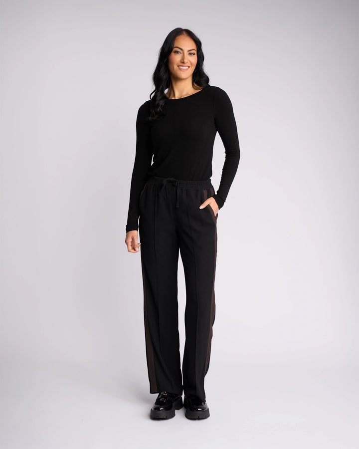 A woman stands against a plain white background, smiling at the camera with her right hand in her pocket. She wears the Knewe Label Bowie Pant in Black/Chocolate, featuring a straight-leg cut and an elasticated waistband.