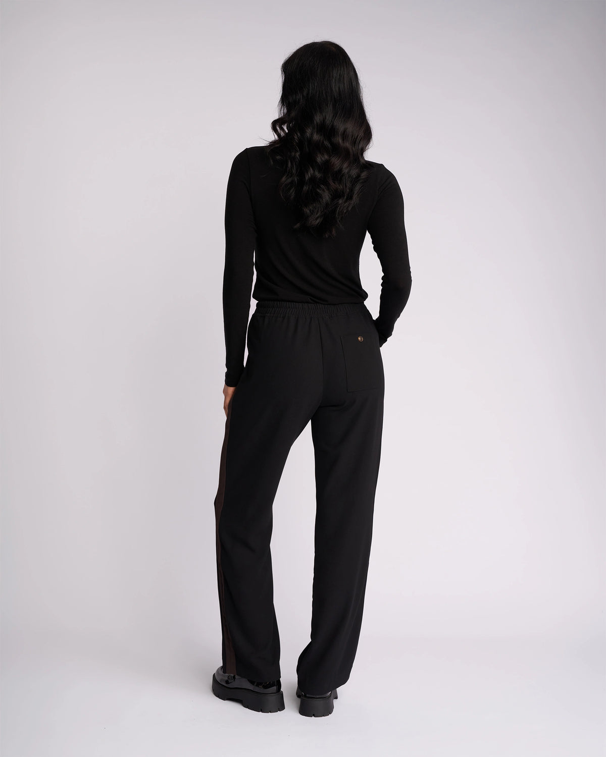 Woman with long wavy dark hair faces away, wearing Knewe Label’s Bowie Pant in Black/Chocolate paired with a black long-sleeve top and black shoes against a plain light background.