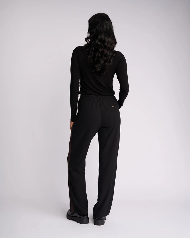 Woman with long wavy dark hair faces away, wearing Knewe Label’s Bowie Pant in Black/Chocolate paired with a black long-sleeve top and black shoes against a plain light background.