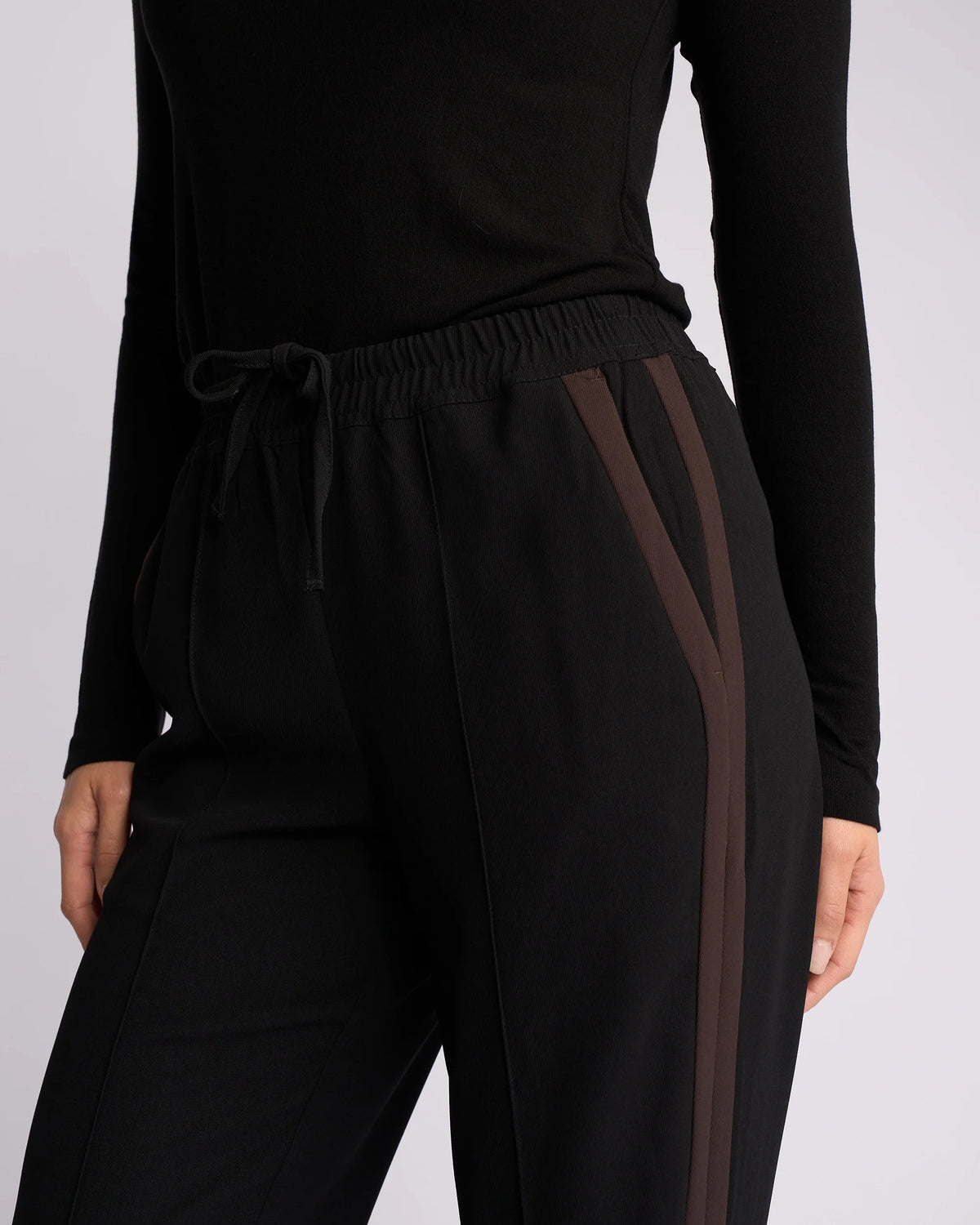 A model wears the Knewe Label Bowie Pant – Black/Chocolate, featuring brown contrast stripes and an elasticated waistband, paired with a black long-sleeve top. Shown from shoulder to mid-thigh against a plain background.