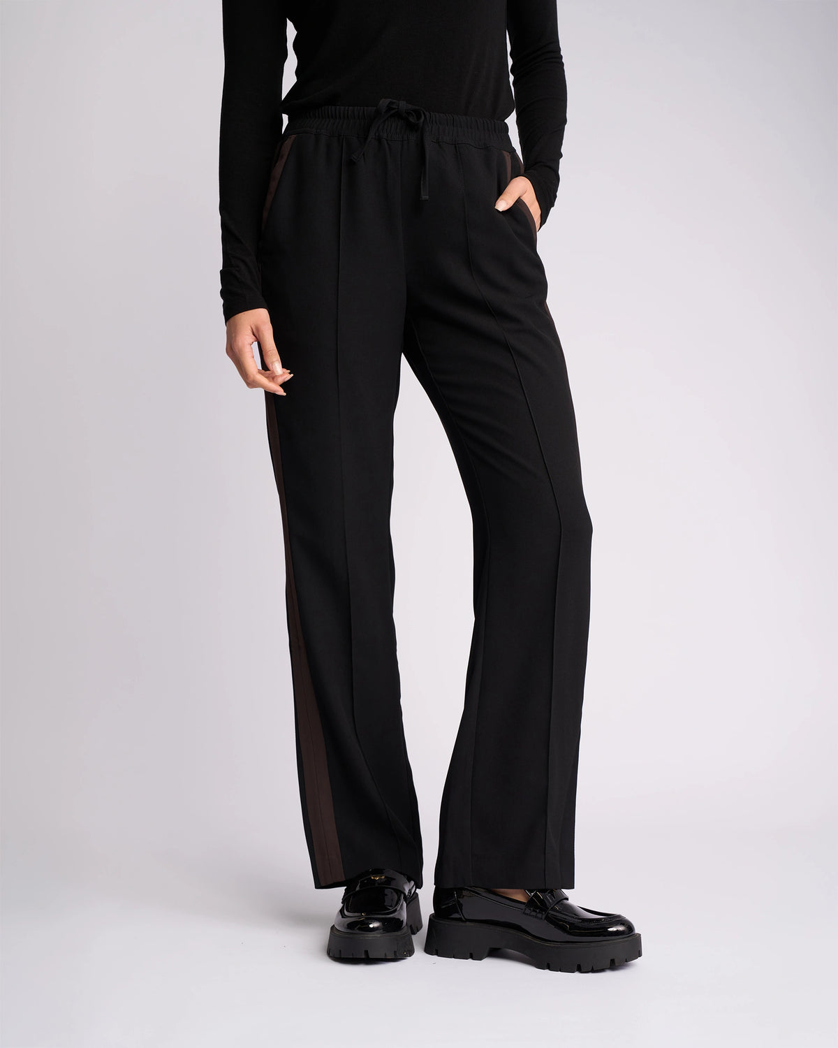 A person wears the Knewe Label Bowie Pant in Black/Chocolate—straight-leg with contrast stripes and elasticated waistband—with a black long-sleeve top and platform shoes, standing against a plain light background, left hand in pocket, head not visible.