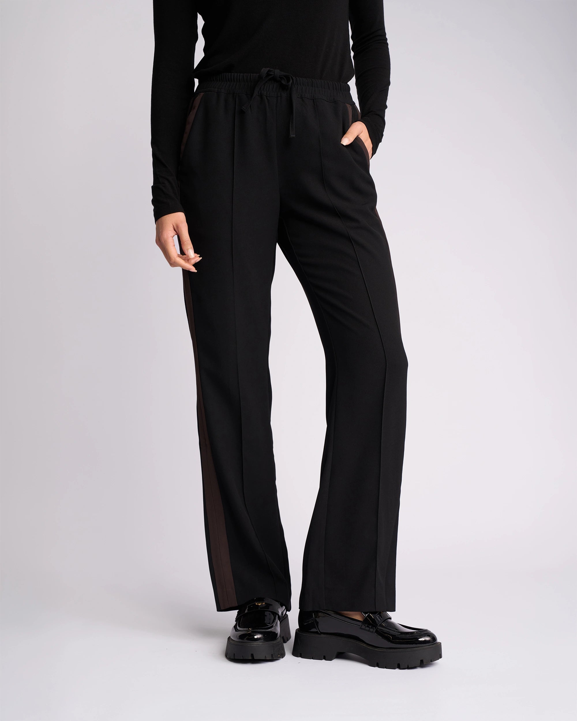 A person wears the Knewe Label Bowie Pant in Black/Chocolate—straight-leg with contrast stripes and elasticated waistband—with a black long-sleeve top and platform shoes, standing against a plain light background, left hand in pocket, head not visible.