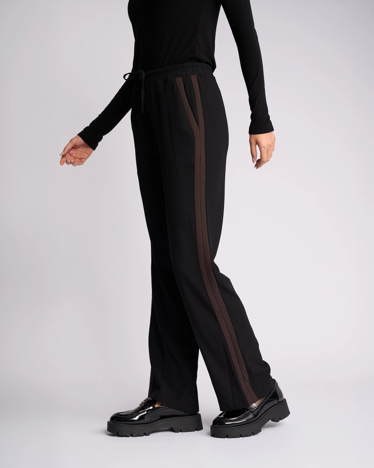Wearing the Knewe Label Bowie Pant in Black/Chocolate, featuring straight legs, contrast stripes, and an elasticated waist, paired with black platform shoes; only the lower half appears against a plain light background.