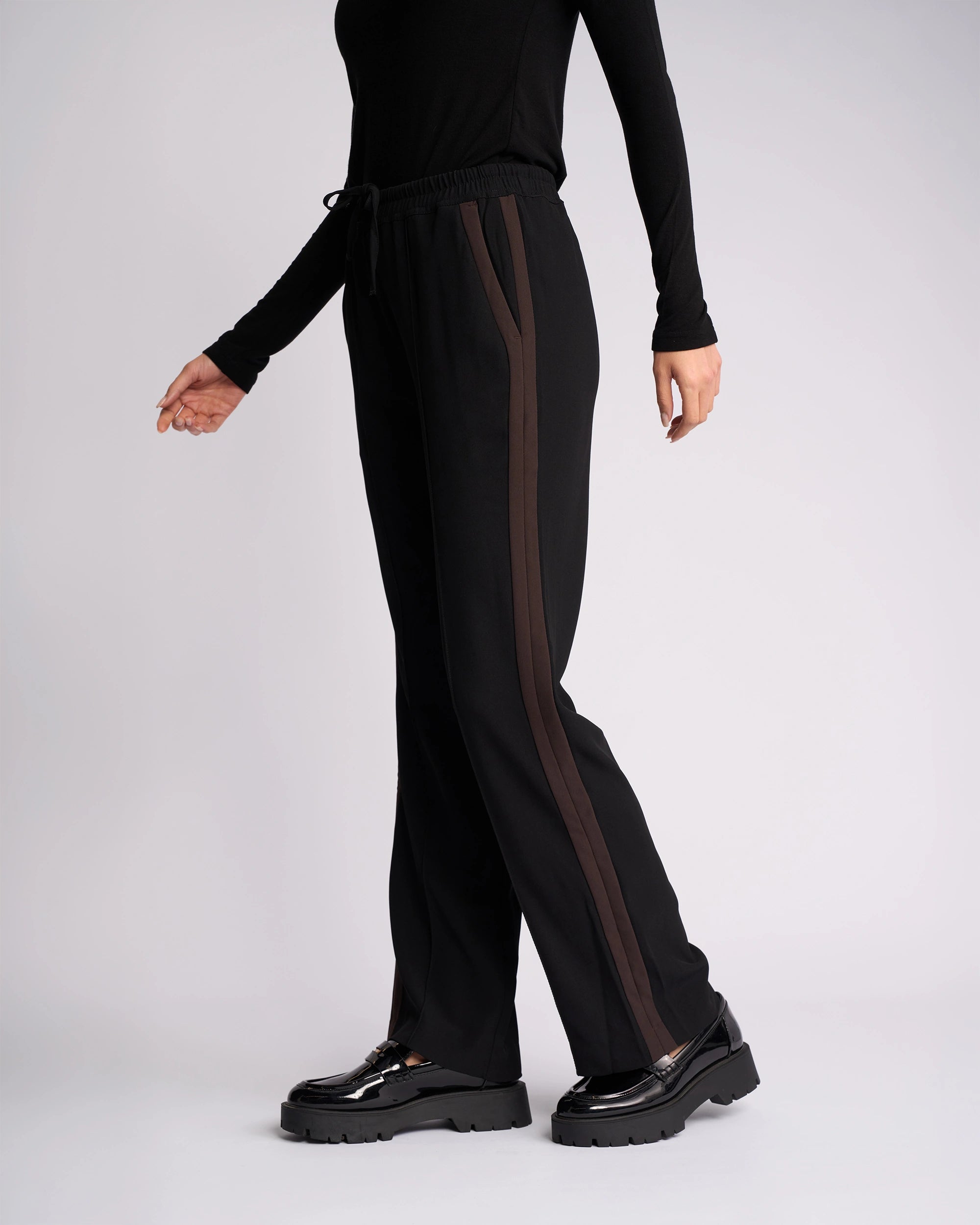 Wearing the Knewe Label Bowie Pant in Black/Chocolate, featuring straight legs, contrast stripes, and an elasticated waist, paired with black platform shoes; only the lower half appears against a plain light background.