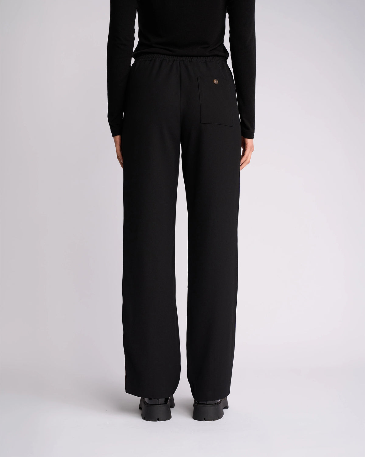 A person faces away from the camera wearing Knewe Label's Bowie Pant in Black/Chocolate—loose-fitting, straight-leg with an elastic waistband and single back pocket—paired with a black long-sleeve top and shoes, set against a white background.