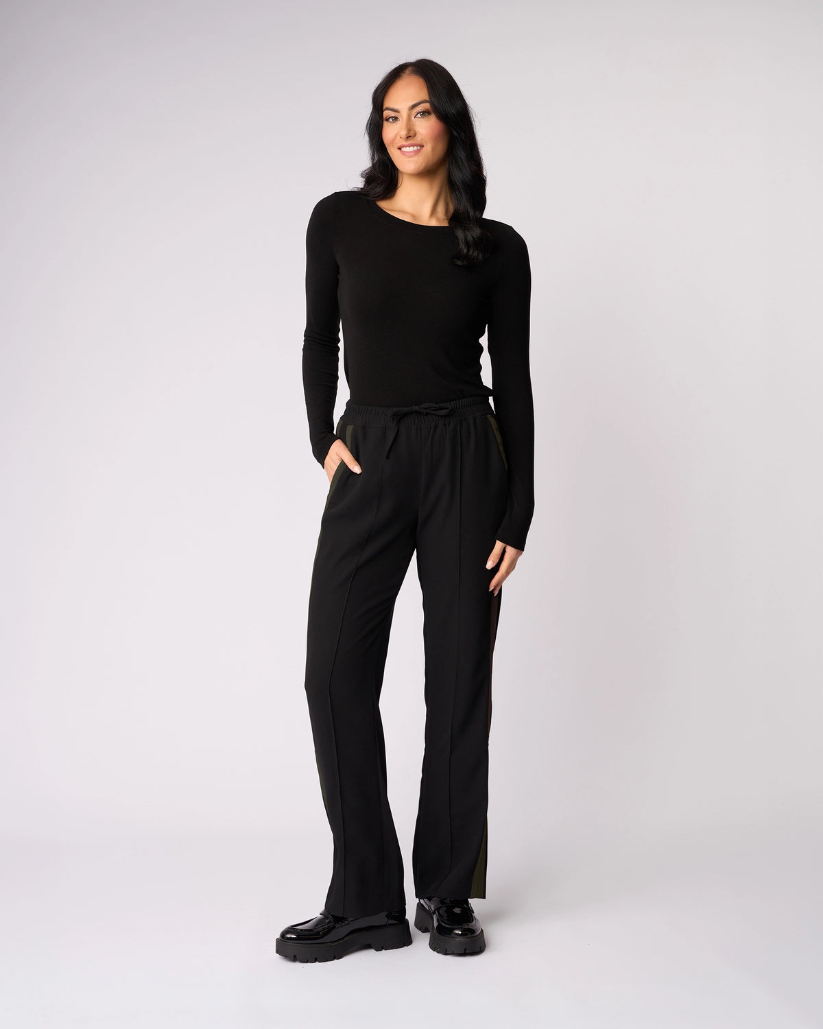 A woman with long dark hair smiles against a plain light background, wearing the Knewe Label Bowie Pant in Black/Moss with an elasticated waistband, a long-sleeve black top, and black shoes. One hand is in her pocket.