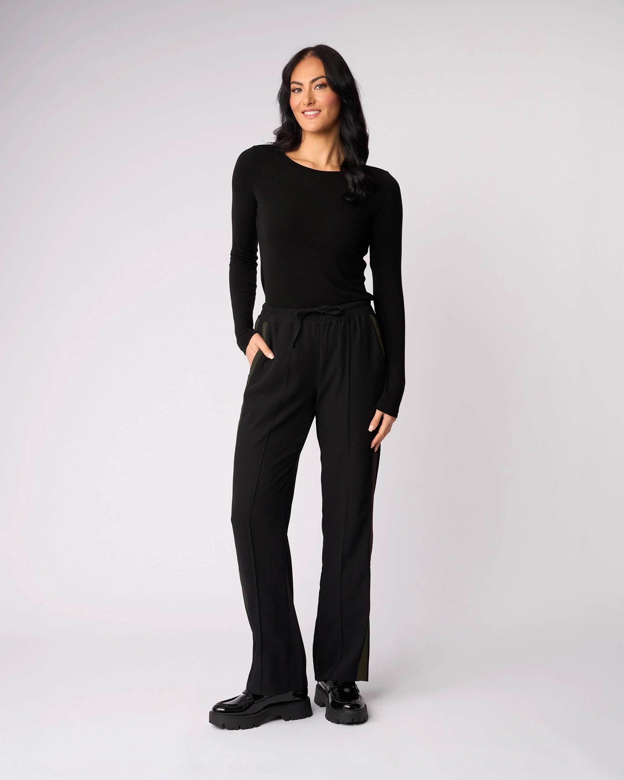 A woman with long dark hair smiles against a plain light background, wearing the Knewe Label Bowie Pant in Black/Moss with an elasticated waistband, a long-sleeve black top, and black shoes. One hand is in her pocket.