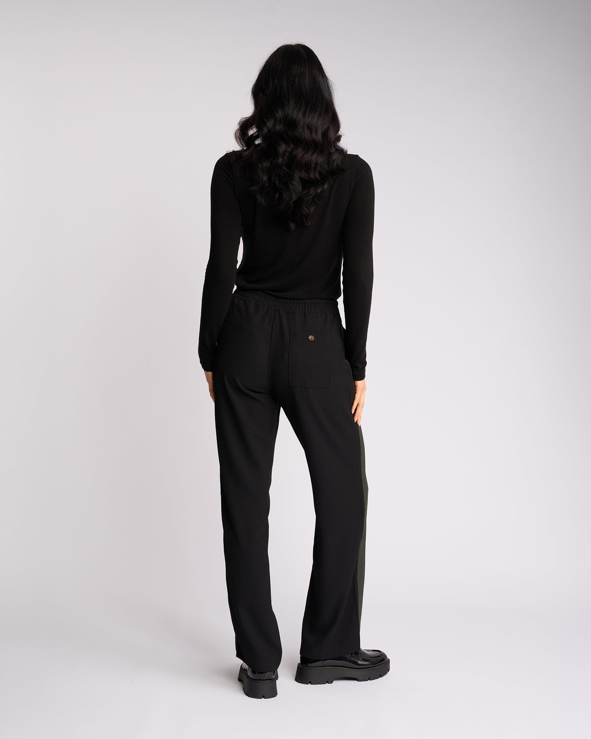 A person with long, wavy dark hair, dressed in a black long-sleeve top and the Knewe Label Bowie Pant - Black/Moss, featuring a pull-on style and elasticated waistband, stands facing away from the camera against a plain white background.