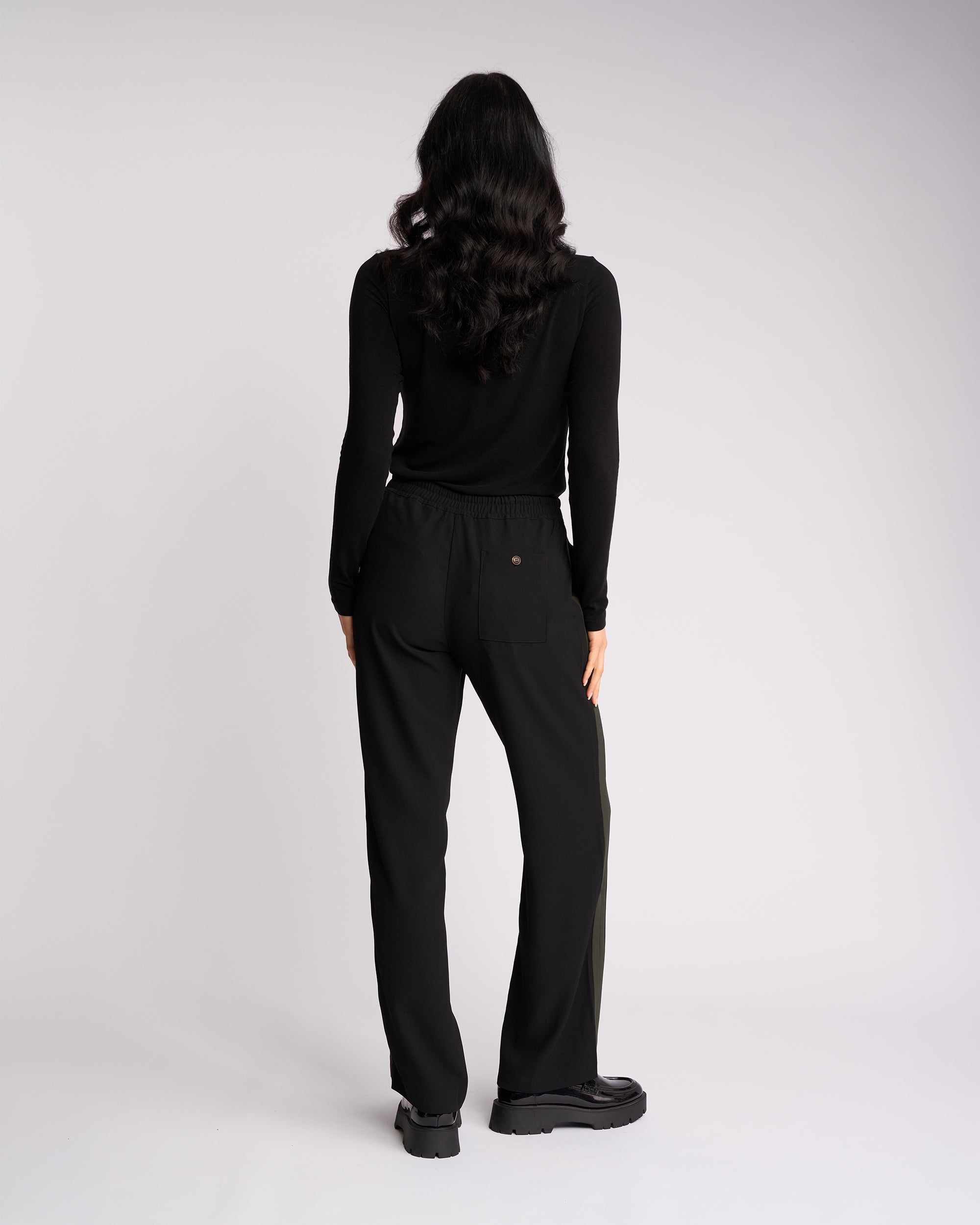 A person with long, wavy dark hair, dressed in a black long-sleeve top and the Knewe Label Bowie Pant - Black/Moss, featuring a pull-on style and elasticated waistband, stands facing away from the camera against a plain white background.